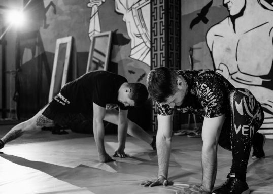 Two men stretching inside a gym, preparing for MMA training in a grayscale setting.